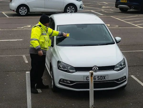 Photo by Caspar Rae on Unsplash - A parking enforcement officer in a high-visibility yellow jacket issuing a ticket to a white Volkswagen car in a parking lot